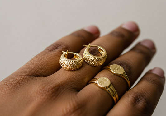 A hand wearing a slim signet ring paired with gold dome hoop earrings.